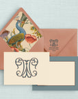 Stationery set with a cream notecard that has an ornate two-letter monogram on the front and blue grey border on the reverse side and a dark salmon colored envelope with a colorful peacock liner and a return address with monogram on the envelope flap.