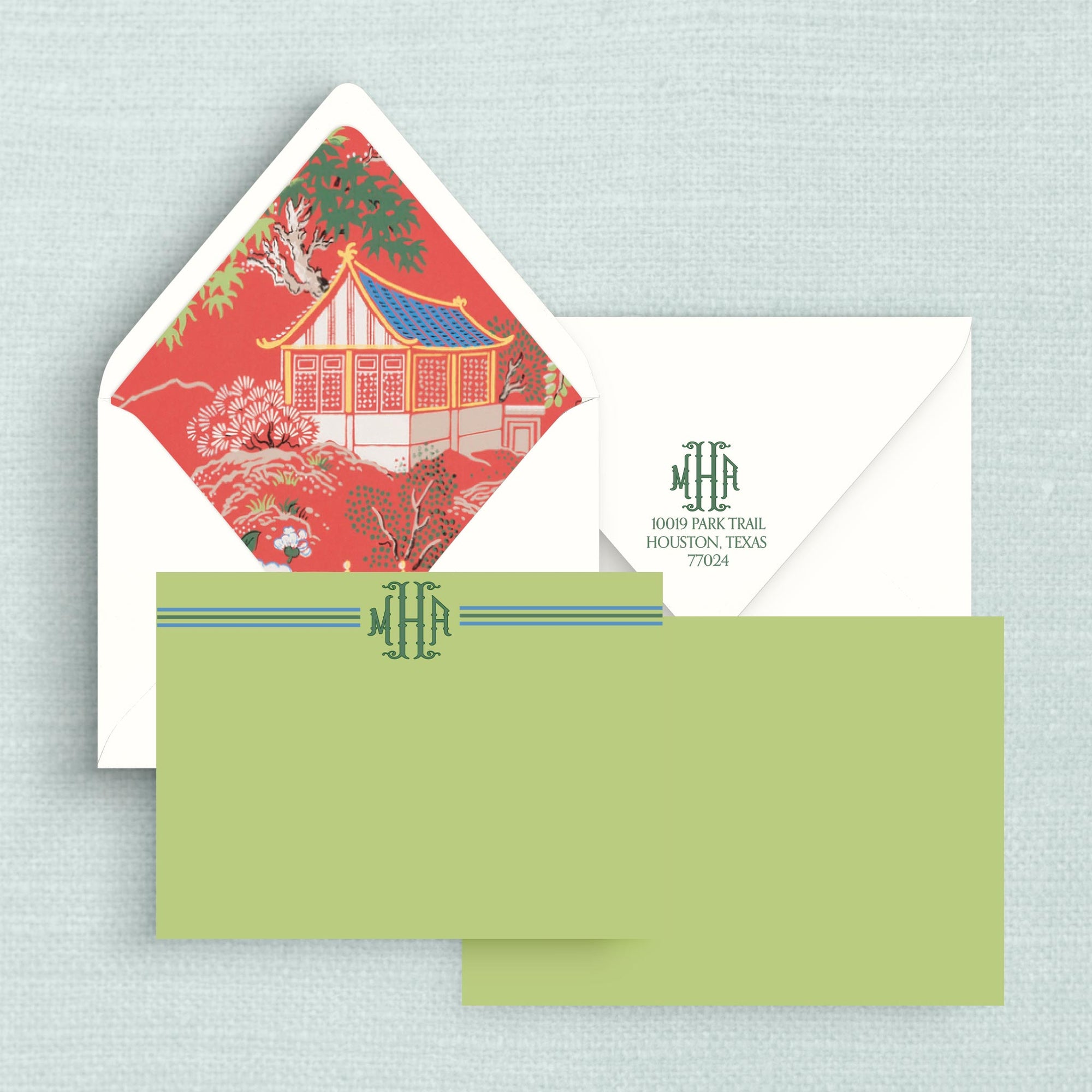 A stationery set featuring a white envelope with return address on flap and a red chinoiserie lining, a two-sided green notecard with monogram lettering on front, all arranged on a pale blue textured background.