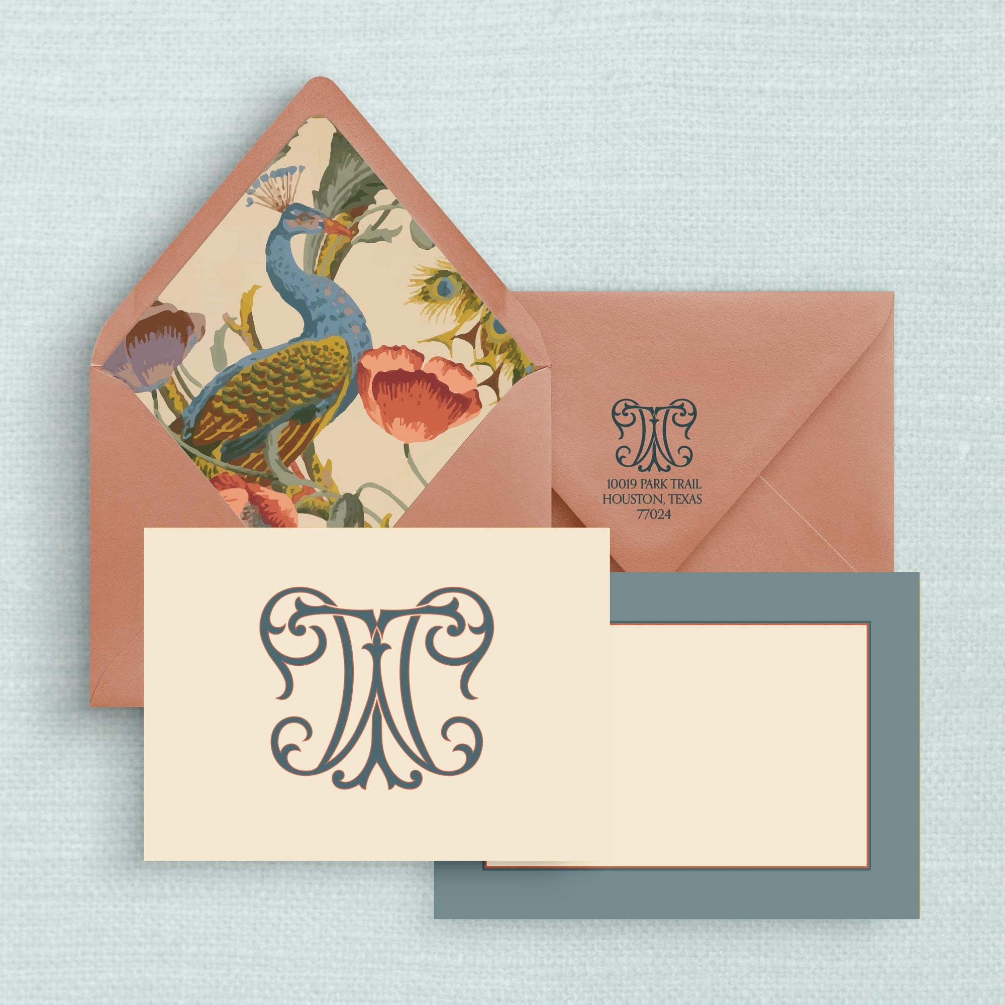 Stationery set with a cream notecard that has an ornate two-letter monogram on the front and blue grey border on the reverse side and a dark salmon colored envelope with a colorful peacock liner and a return address with monogram on the envelope flap.