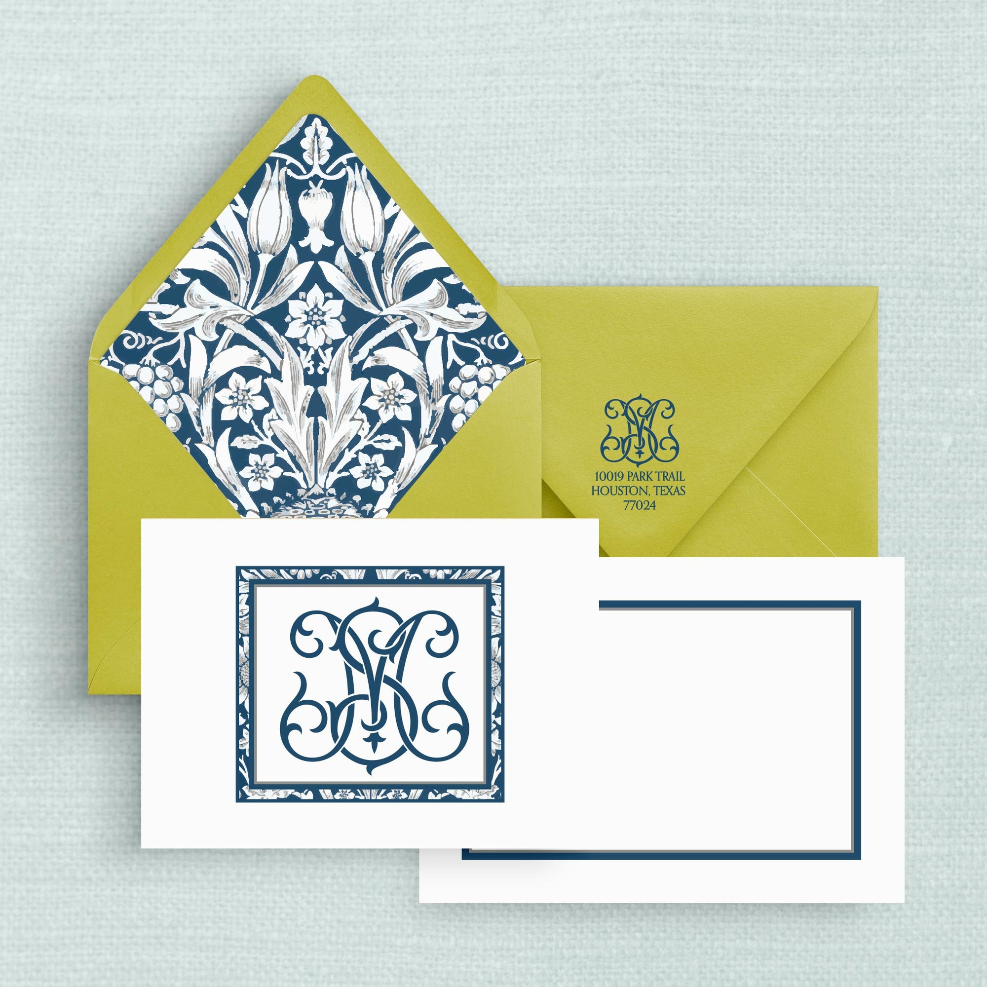 A stationery set featuring a two-sided note card in white with a navy blue ornate two-letter monogram and border and simple border on the reverse side, a matching bright green envelope with a blue and white floral patterned liner and return address with monogram on the flap.